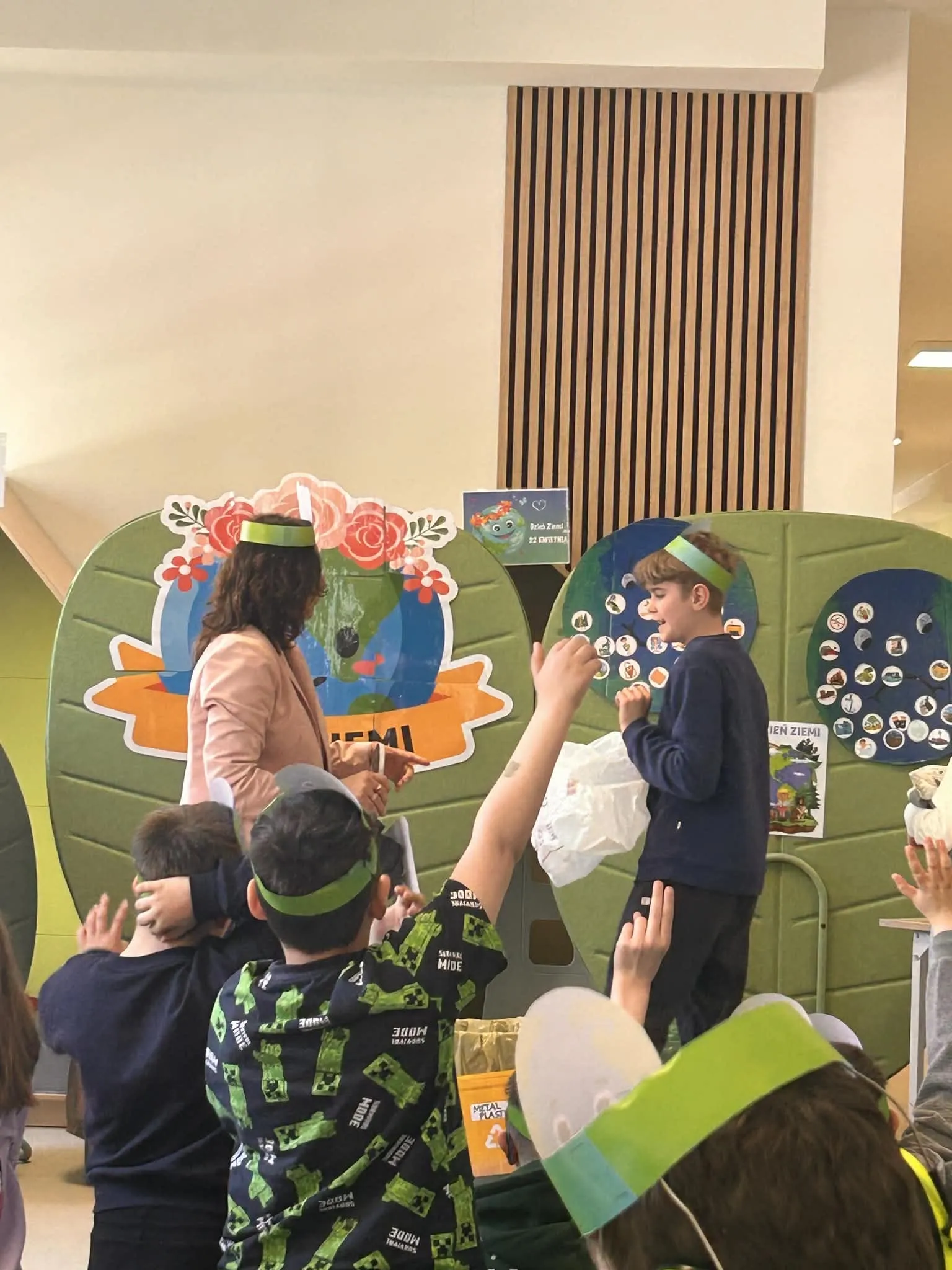 Na zdjęciu uczeń oraz nauczycielka na korytarzu szkolnym - stoją przy planszach  z  różnymi  elementami oraz  plakacie z kulą ziemską. Oboje mają zielone opaski na głowie.  W dolnej części zdjęcia widoczni są inni uczniowie.