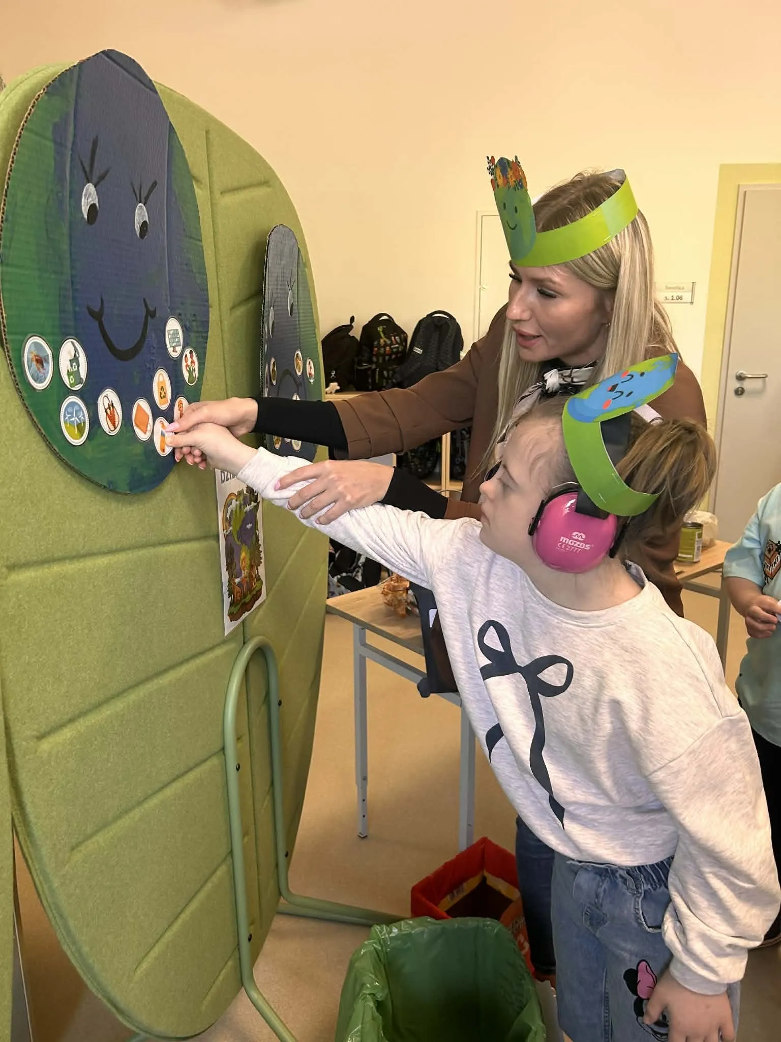 Na zdjęciu uczennica z nauczycielką - obie stoją przed planszą z różnymi elementami, uczennica z pomocą nauczycielki  przesuwa lub nakłada jakiś element na planszę. 