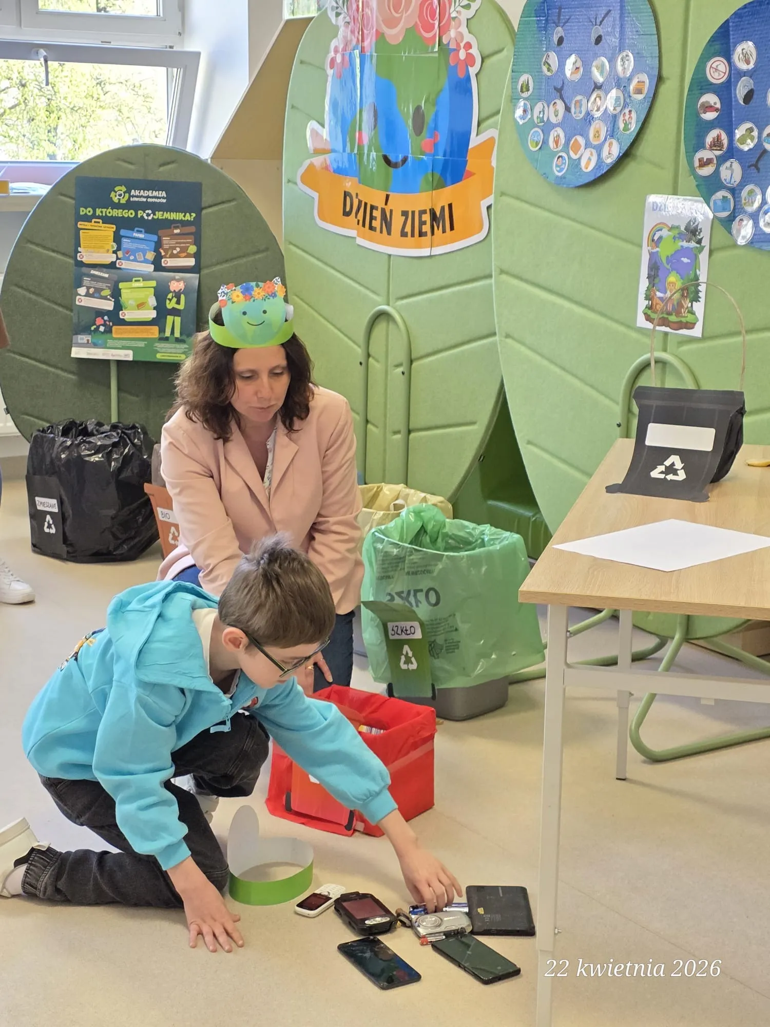 Na zdjęciu uczeń, i nauczycielka na korytarzu szkolnym. Uczeń pochyla się przy leżących na podłodze gadżetach elektronicznych. Obok widoczne są kosze na odpady w zielonym i czerwonym, żółtym  oraz dalej czarnym kolorze. W tle plansze związane z ćwiczeniami edukacyjnymi  dotyczącymi ochrony Ziemi.