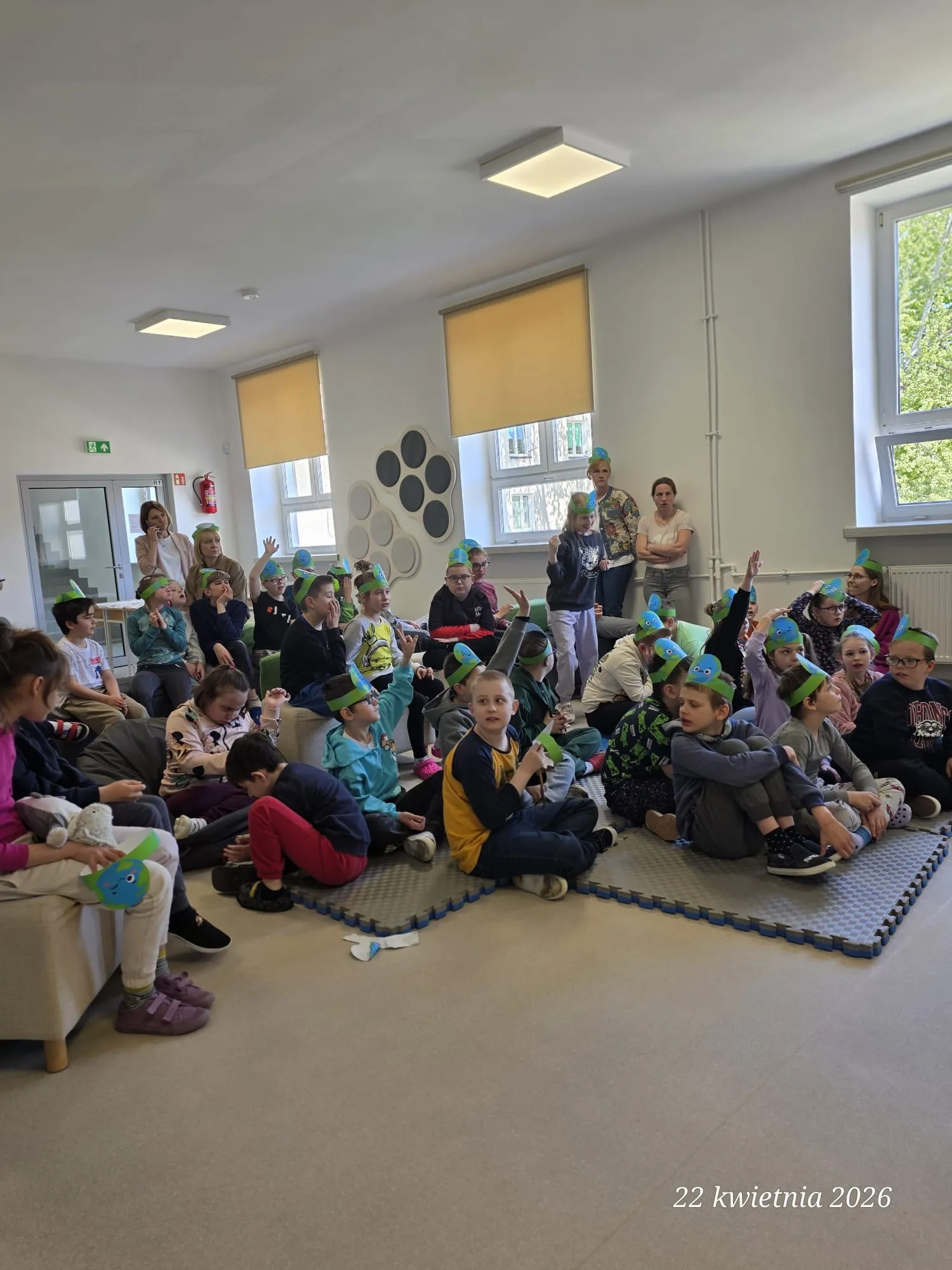 Na korytarzu szkolnym siedzą  na matach uczniowie  - w czapeczkach  z wizerunkiem Ziemi. W głębi zdjęcia widoczni są nauczyciele i pomocnicy.