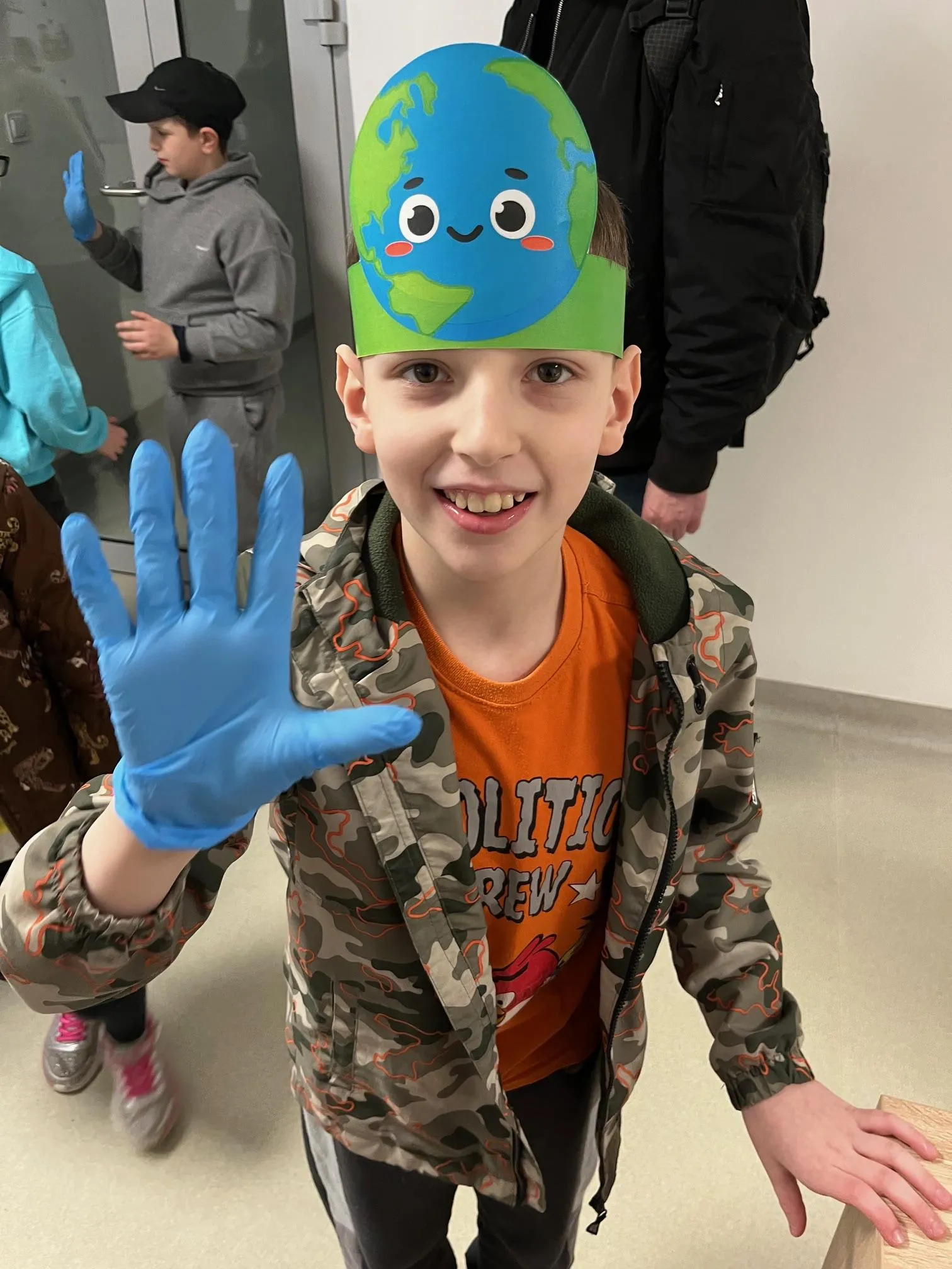 Na zdjęciu uczeń w czapeczce z wizerunkiem Ziemi, pokazujący rękę w  ochronnej rękawiczce. W głębi zdjecia widoczni są inni uczniowie.