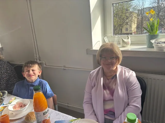 Na pierwszym planie, przy świątecznym stole , znajdującym się na korytarzu szkolnym siedzi  nauczycielka rytmiki i jeden z uczniów.