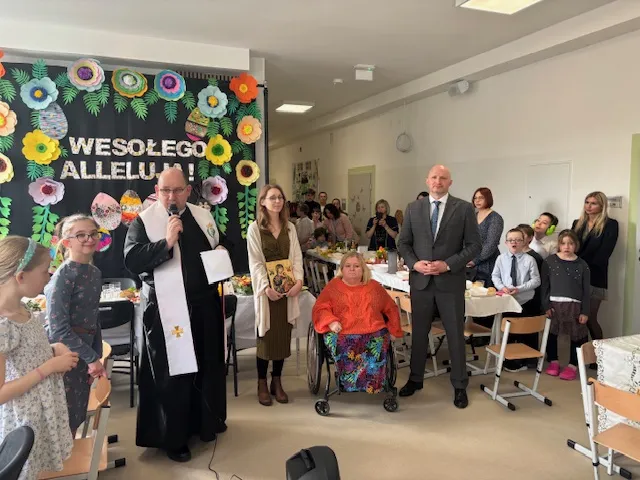 W tle tablicy z dekoracja wielkanocną stoi na korytarzu szkolnym ksiądz z mikrofonem w ręku, obok wicedyrektorka szkoły przy ulicy Żytniej oraz dyrektor szkoły nr 147 i pani pedagog. Z prawej strony księdza stoi jedna z uczennic szkoły, a   w dalszej części zdjęcia widoczny jest zastawiony świąteczny stół oraz uczniowie i nauczyciele stojący wokół stołu.