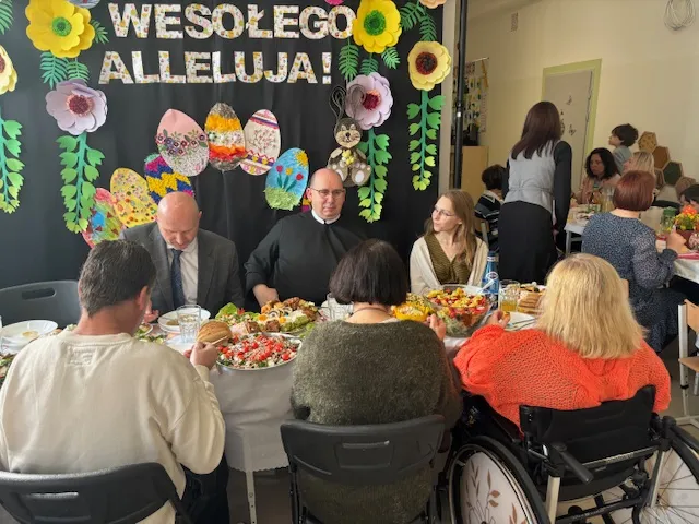 Na pierwszym planie, przy zastawionym świątecznym stole , znajdującym się na korytarzu szkolnym siedzi ksiądz, po jego obu stronach dyrektor i wicedyrektorka szkoły oraz na przeciwko nich  - inne trzy osoby dorosłe. Wyżej ponad stołem widać tablice z dekoracją świąteczną. Dalej widoczny jest fragment innego świątecznego stołu z uczniami i  osobami dorosłymi. 