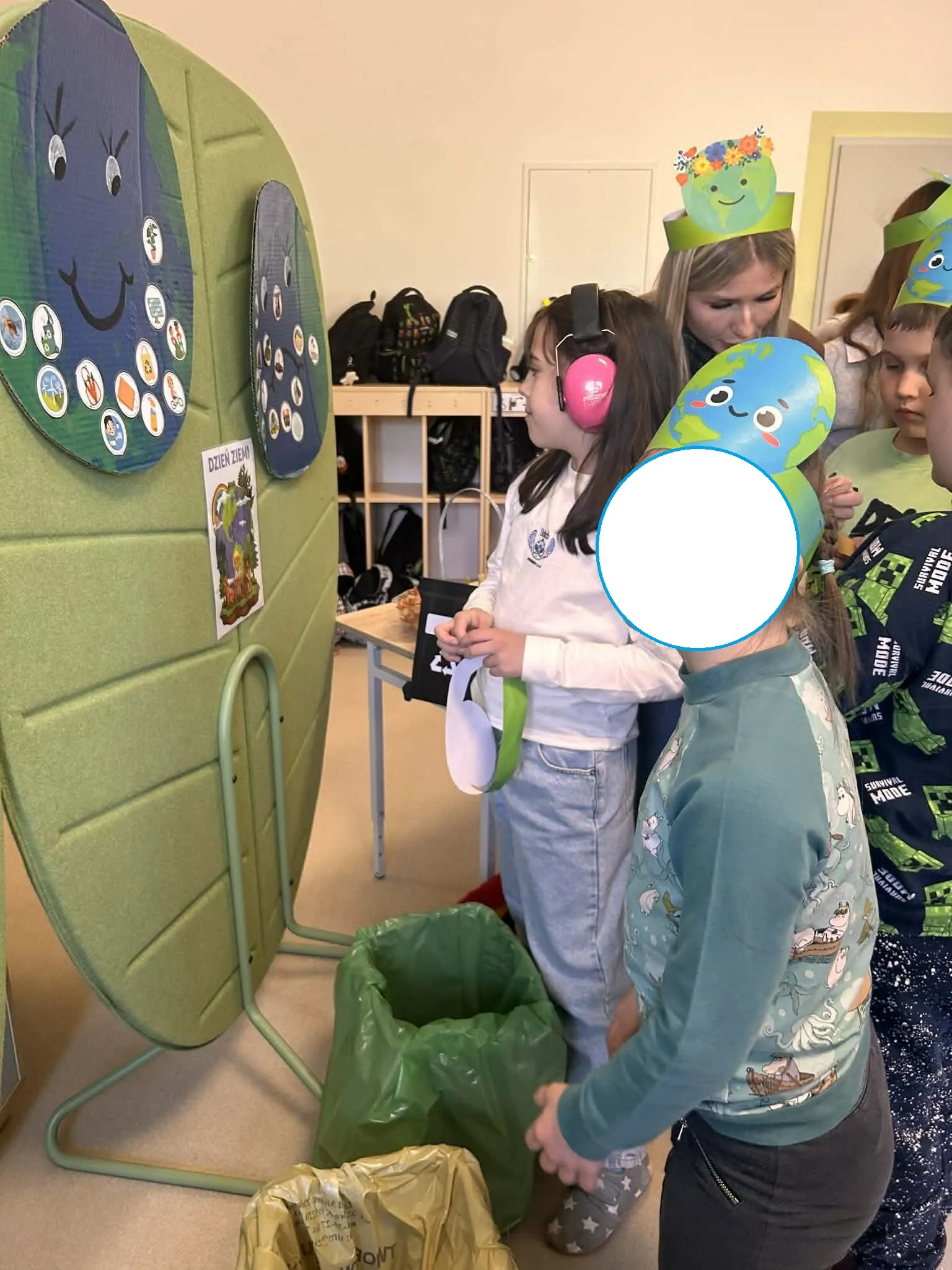 Uczniowie stoją na korytarzu szkolnym przed planszami z uśmiechniętą i smutną ziemią i przyklejają do tych plansz odpowiednie obrazki .  Obok uczniów stoi nauczycielka.