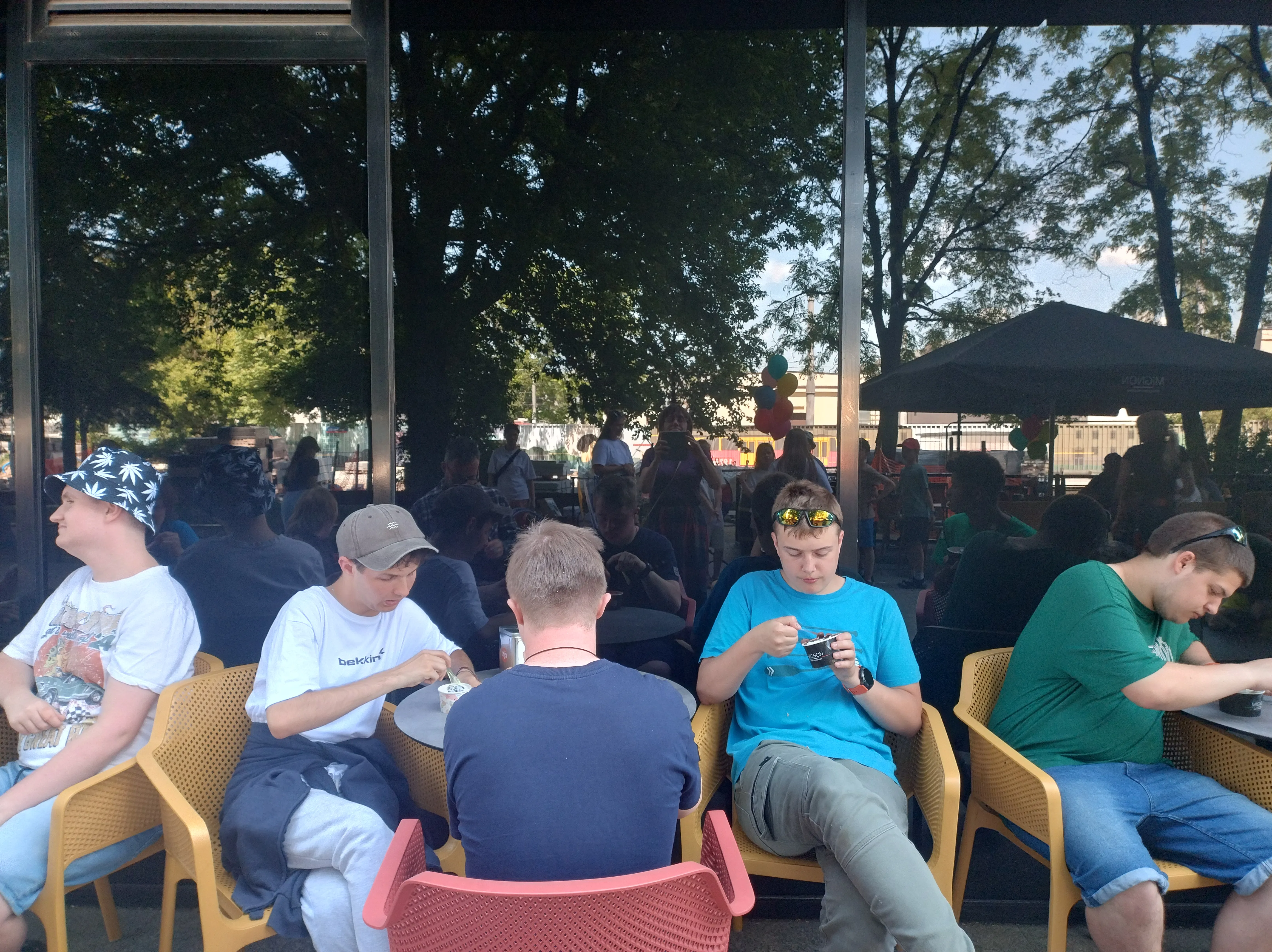 Na zdjęciu uczniowie siedzący przy stolikach przed barem restauracyjnym, jedzący lody.