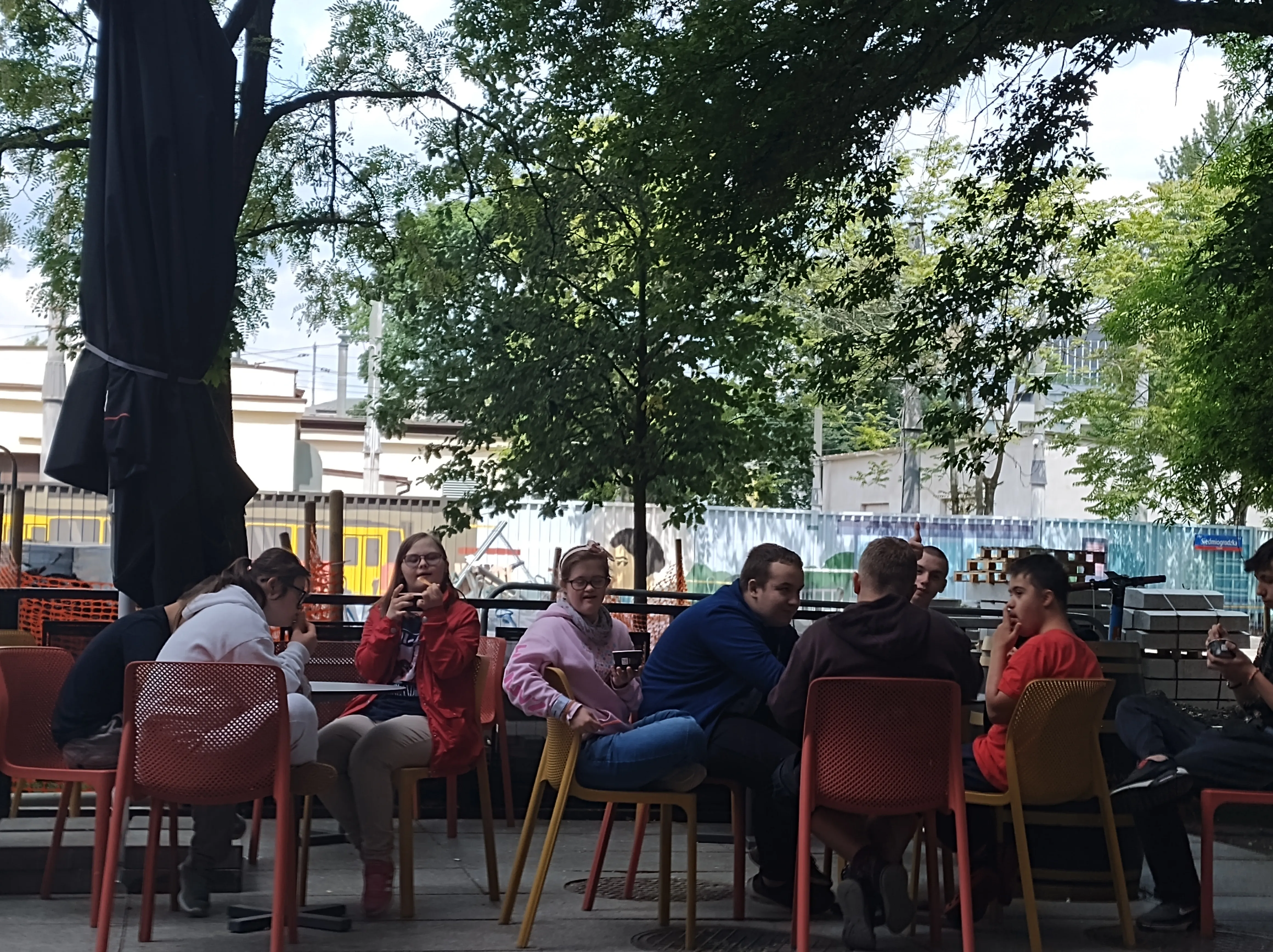 Na zdjęciu uczniowie, którzy siedzą  przy stolikach przed barem restauracyjnym i jedzą lody. 
