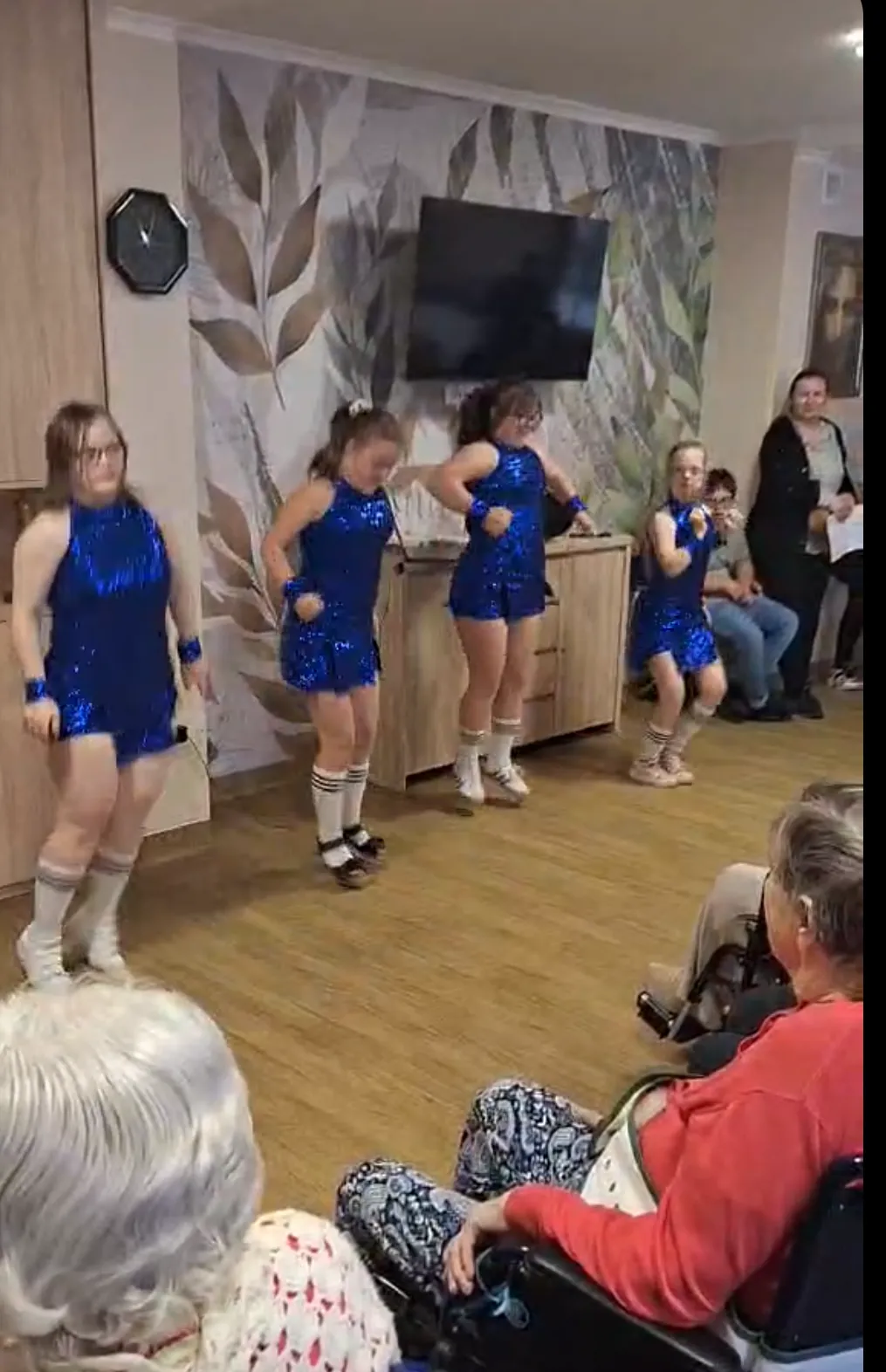 Występ zespołu tanecznego przed pensjonariuszami Zakładu Opiekuńczego. Na zdjęciu widoczne są 4  uczennice w niebieskich strojach tańczące na scenie przed publicznością. 