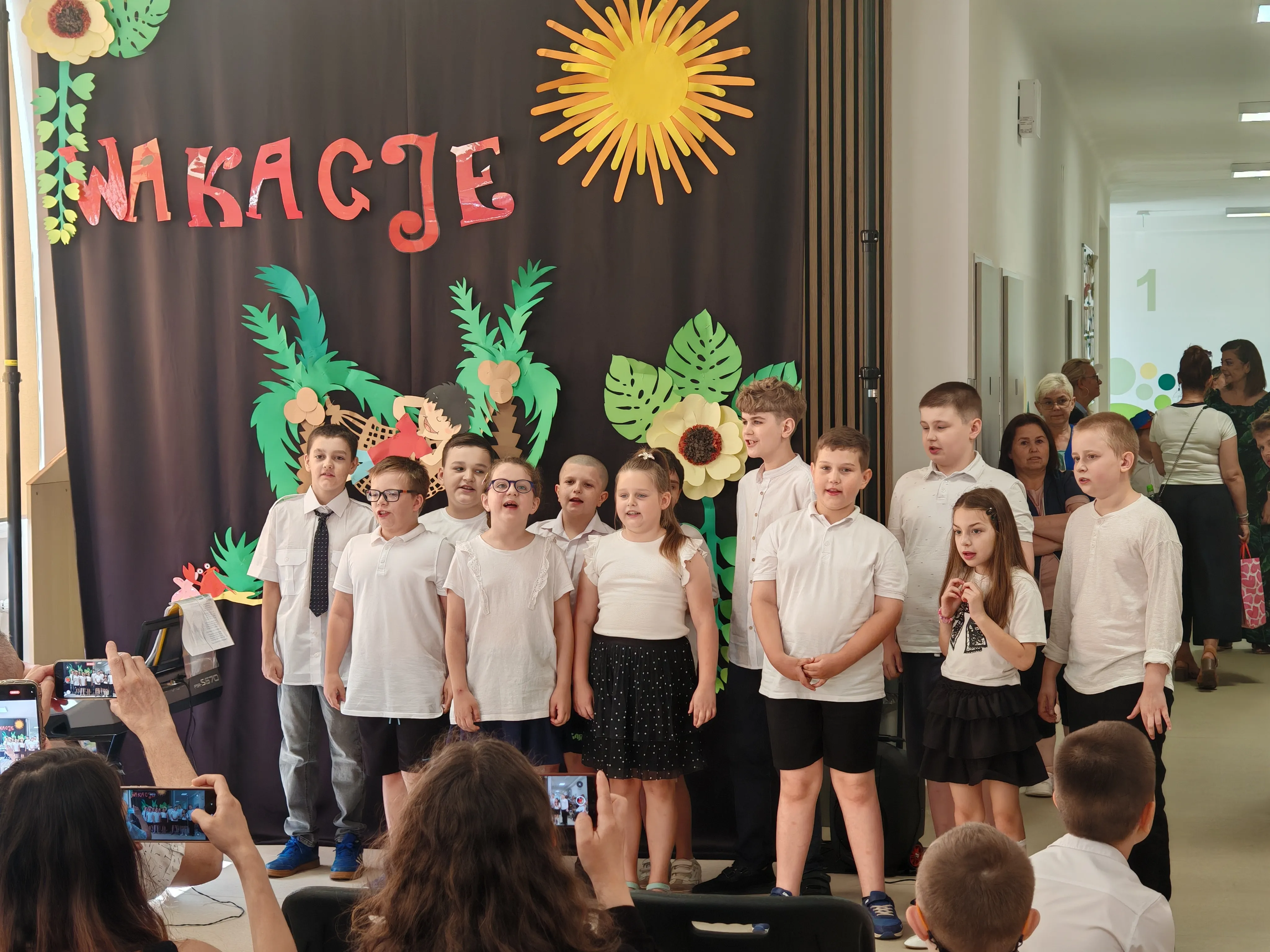 Występ  dzieci z klas 1-3, w galowych strojach stoją przed widownią na korytarzu szkolnym na tle wakacyjnej dekoracji.