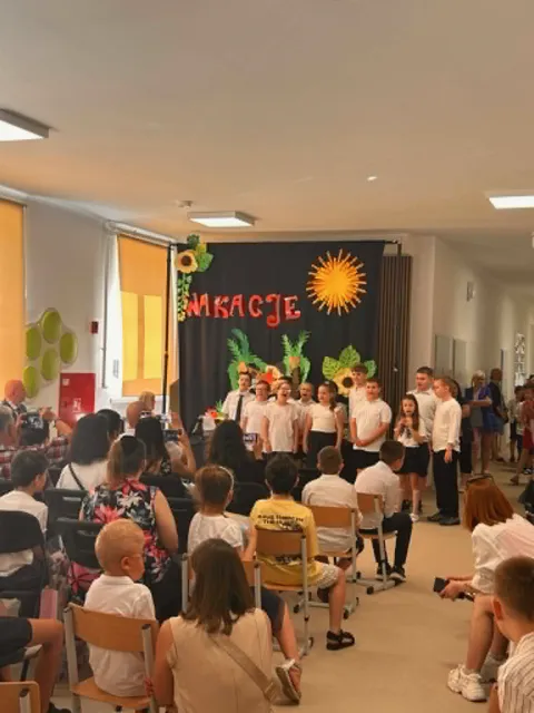 Występ  dzieci z klas 1-3, w galowych strojach stoją przed widownią na korytarzu szkolnym na tle wakacyjnej dekoracji.