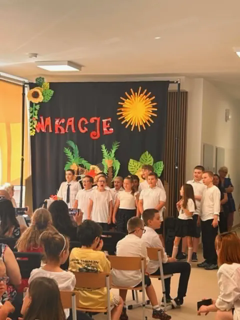 Występ  dzieci z klas 1-3, w galowych strojach stoją przed widownią na korytarzu szkolnym na tle wakacyjnej dekoracji.